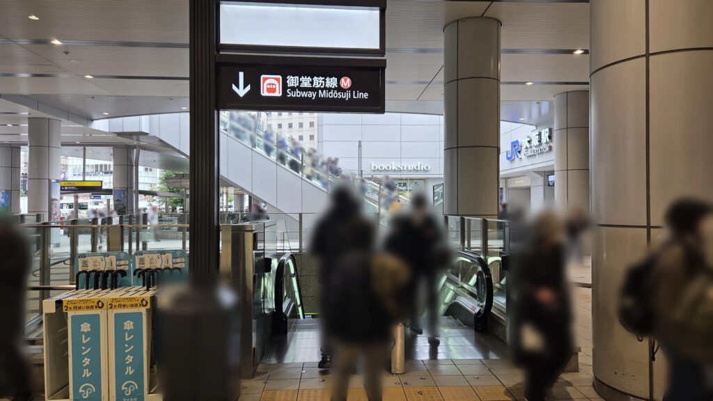 地下へ続くエスカレーターの様子。頭上には御堂筋線を示すアイコンと矢印が書かれた看板があり、正面奥には「bookstudio」の文字が見えます。