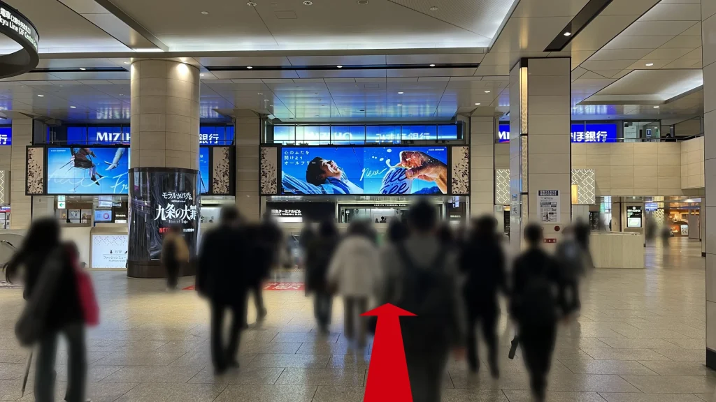 Alt：Signatureグラングリーンゲートタワーへのアクセス方法：エスカレーターを下りた先の1Fコンコースで、正面に「みずほ銀行」の看板や大型のデジタルサイネージがあり、多くの人々が歩いている様子。