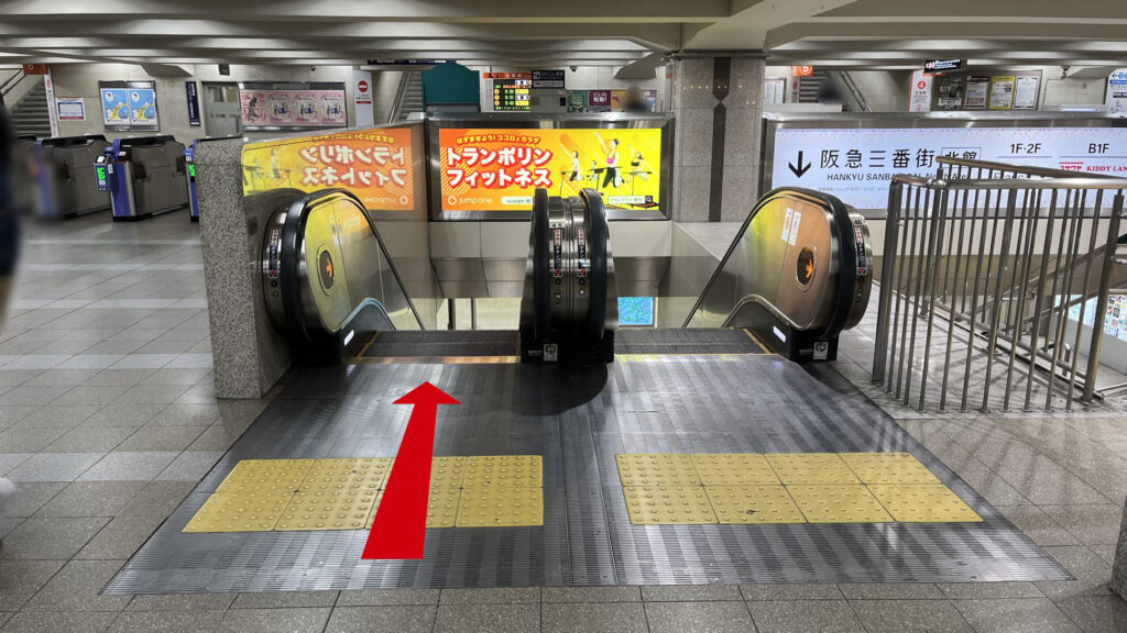 billage OSAKA 朝日プラザ梅田へのアクセス：1Fへ降りるための下りエスカレーター　左正面に改札、エスカレーター上部にトランポリンフィットネスの広告がある