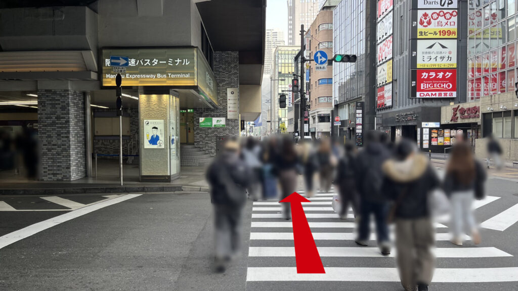 billage OSAKA 朝日プラザ梅田へのアクセス：屋外へ出て信号を渡る様子　向かいに「阪急バスターミナル」の看板と銀行ATM、反対側の歩道沿いには居酒屋の看板等が見える