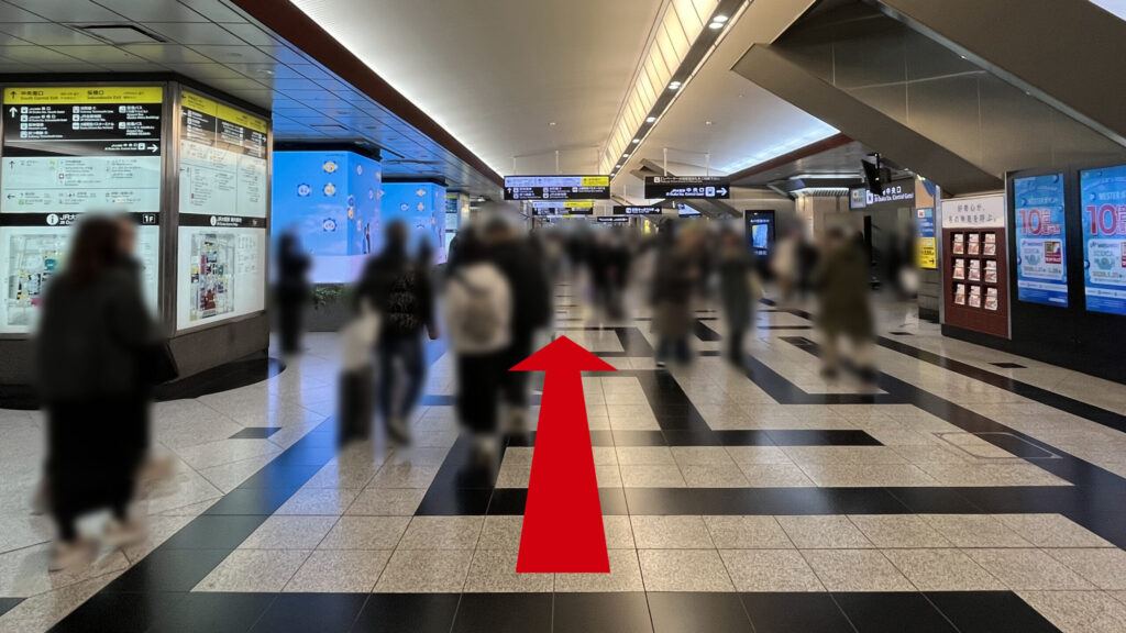 リージャス梅田スクエアビジネスセンターへのアクセス方法：白と黒の幾何学模様の床が広がるコンコースを進む様子　左右にはデジタルサイネージや大きな柱が立っている