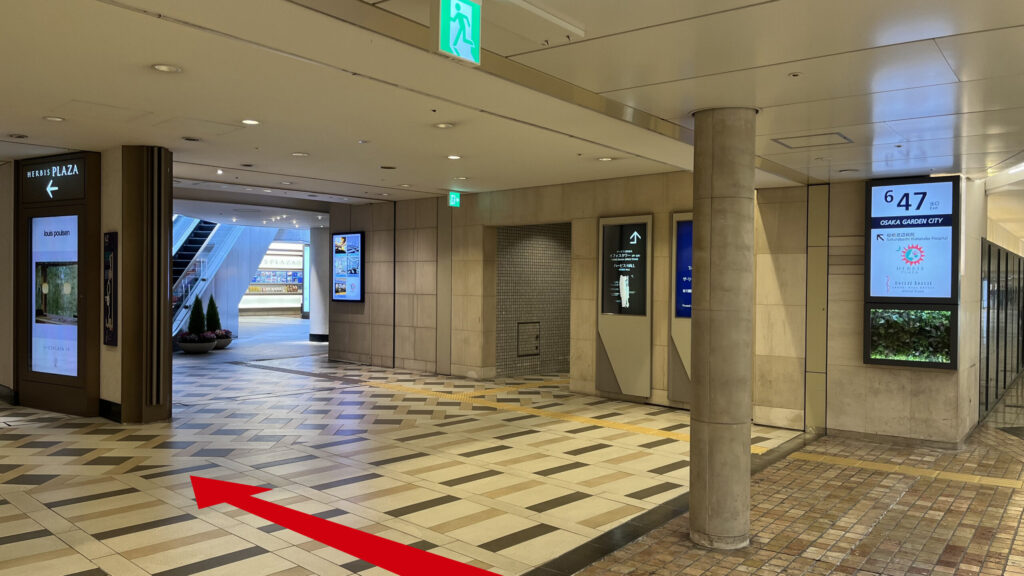 SYNTH×Business-Airport 西梅田ブリーゼタワーへのアクセス方法：地下通路「大阪ガーデンシティ」の分岐点。右側の柱にあるデジタルサイネージに「6-47」の出口番号と「OSAKA GARDEN CITY」「HERBIS PLAZA」の文字、左方向へ「BREEZE TOWER」を指す白い看板が設置されている様子。