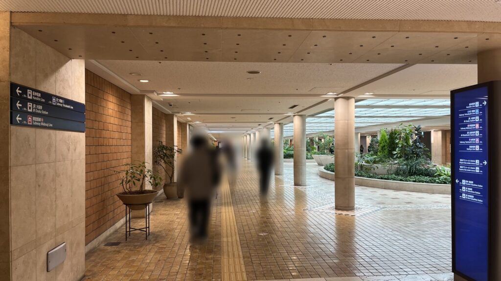 リージャスパシフィックマークス西梅田ビジネスセンターへのアクセス方法：左右に路線案内板がある通路　白い円柱が並んでいるが、その右手に吹き抜けと緑の生い茂った噴水がある