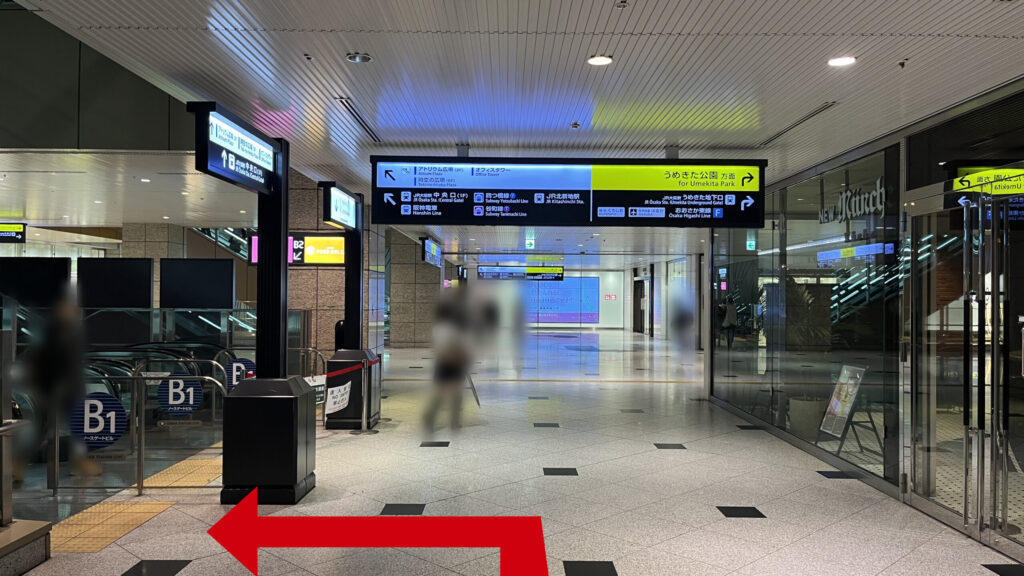 Compass Offices イノゲート大阪へのアクセス方法：上りエスカレーターの乗り口。右側には「ニューミュンヘン」の店舗、頭上には「うめきた公園方面」を示す黄色い案内板がありその手前で左に曲がる