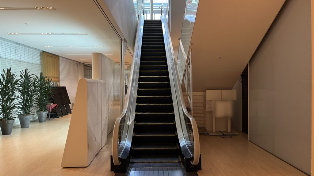 SYNTH×Business-Airport 西梅田ブリーゼタワーへのアクセス方法：1Fフロアへと続くエスカレーターの正面。左側には観葉植物が並び、上部からは1Fフロアの明るい光が差し込んでいる空間。