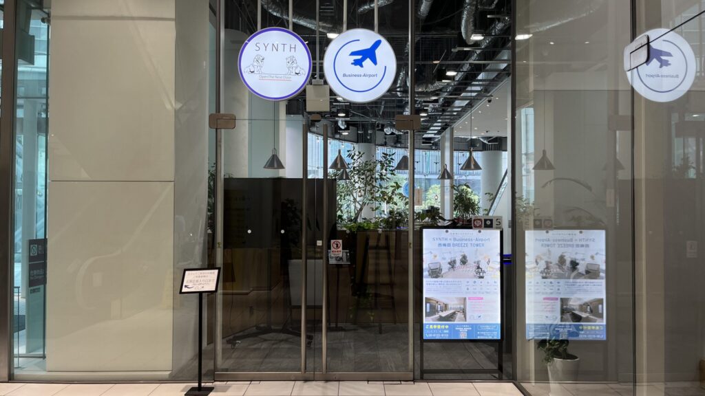 SYNTH×Business-Airport 西梅田ブリーゼタワーへのアクセス方法：1Fフロアのエスカレーター正面にある店舗入り口。ガラス張りの外装上部に「SYNTH」の円形の紫色ロゴと、飛行機マークが入った「Business-Airport」の円形青色ロゴ看板が掲出されている。