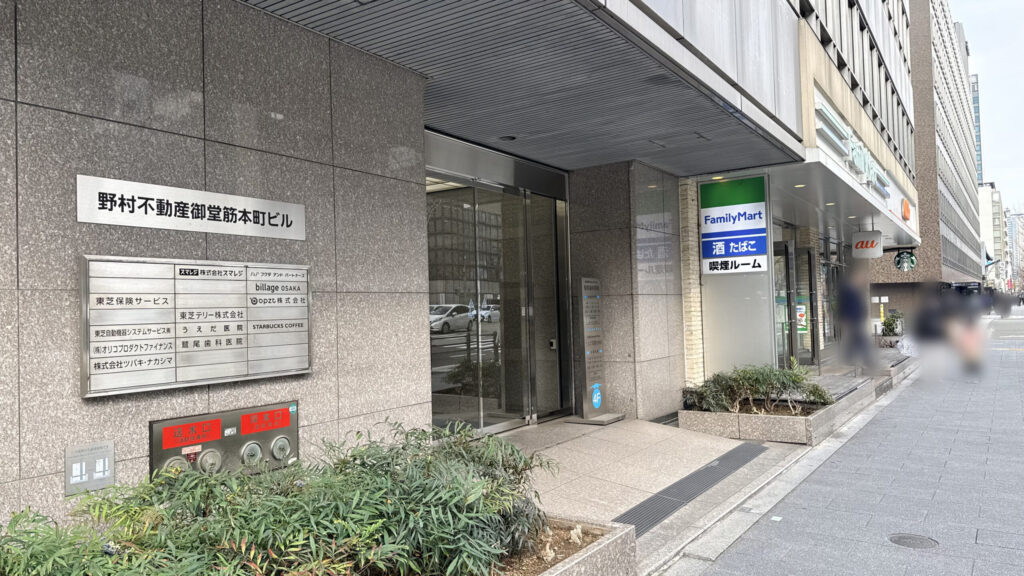 H¹O 本町Ⅱへの行き方:野村不動産御堂筋本町ビルのエントランス。ガラス張りの自動ドアがあり、左側の壁に「H¹O 本町Ⅱ」の黒い看板が設置されている。右隣にファミリーマートがある