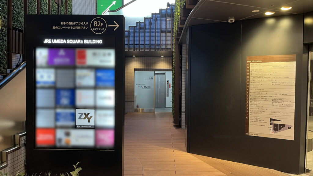 ZXY梅田の入っているJRE梅田スクエアビルのB2連絡通路付近　左にテナント看板があり、その後ろに吹き抜けの階段がある　階段の下にエレベーターがある