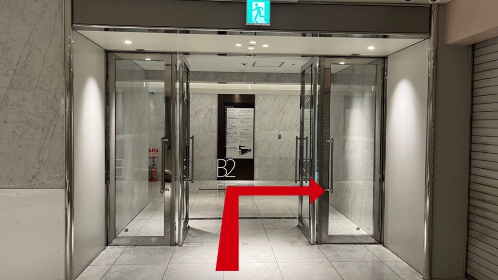 エレベーターホールへ向かう道　ガラス扉があり、奥にB2と書かれたビルの各階テナント一覧が掲示されている