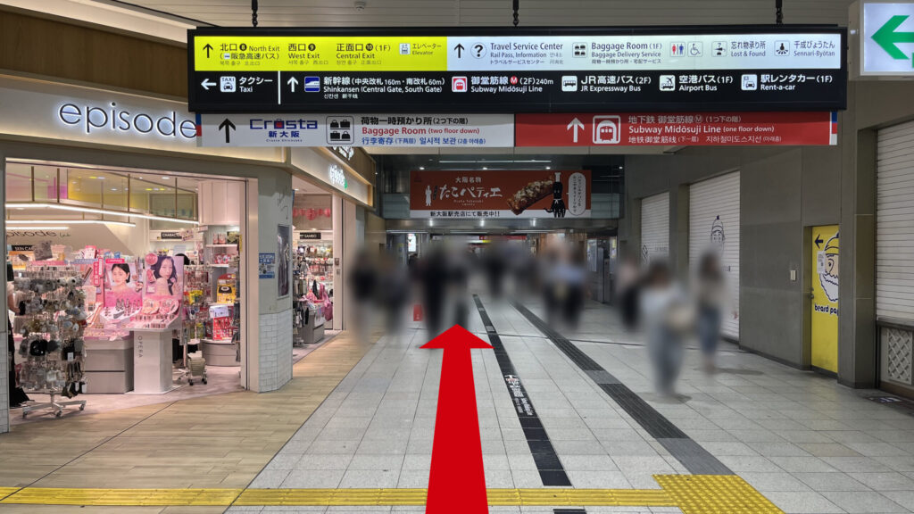 リージャス新大阪阪急ビルビジネスセンターへの行き方:店舗が並ぶ駅構内の広い通路。頭上には各方面への黄色と白の案内看板が設置されており、奥へと続く道を歩行者が進んでいる。