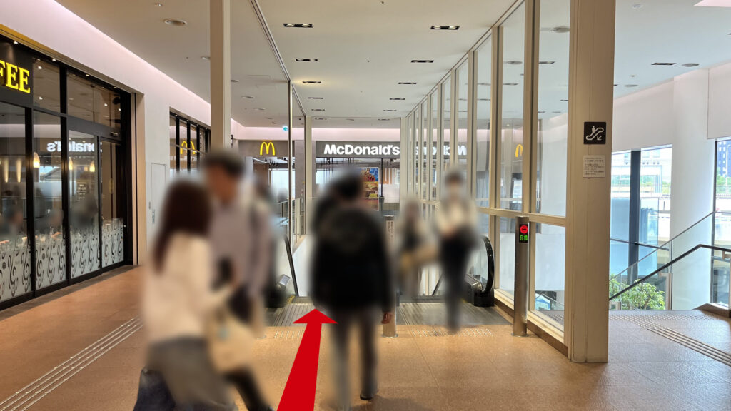 通路の先にあるマクドナルドの店舗の手前で、2Fへとつながる下りエスカレーターと階段の降り口の様子。奥にはエスカレーターを囲うように通っている通路とマクドナルドがある
