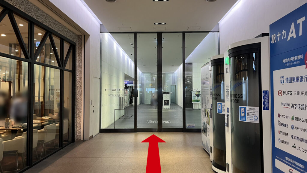 リージャス新大阪阪急ビルビジネスセンターへの行き方:通路の奥にあるガラス張りのホテルのエントランスドアと、手前右側に駅中リモートブースと駅ナカATMの端末が並んでいる