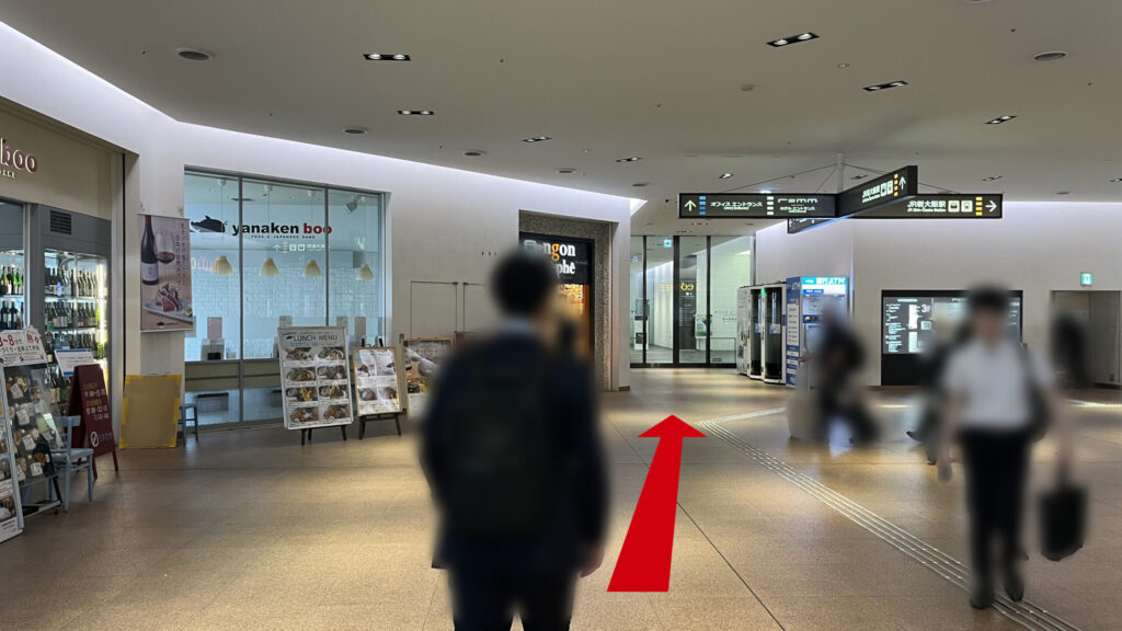 エスカレーターを上りきった先にある広い屋内通路。左側には飲食店のガラス壁が見え、正面奥にはガラス扉のエントランスが視認できる。