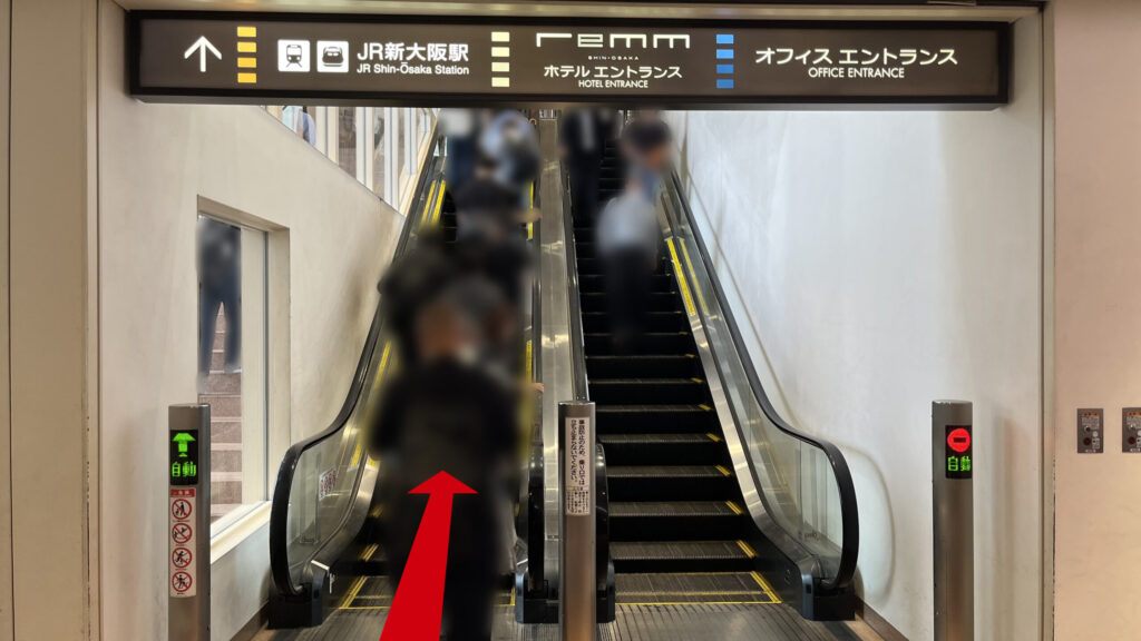 ビル上層階へ続く上りエスカレーターの乗り口。頭上にはJR新大阪駅やオフィスエントランスへと導く黒い案内板が掲げられている。