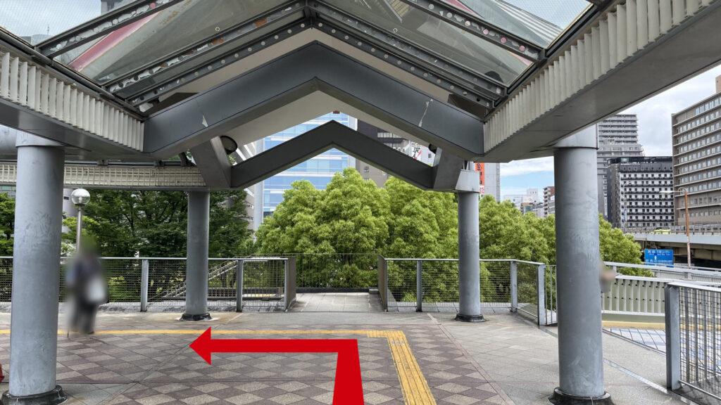 ガラス屋根のある連絡橋の突き当たり部分で、床面には黄色の点字ブロックが敷かれており、前方に緑の木々が見える景色の様子。