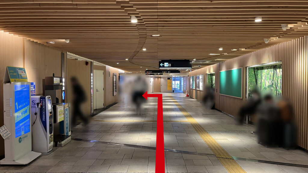 通路の突き当たり付近で、頭上にトイレや出口の案内看板がある 右の壁沿いにはベンチがあり、左の壁沿いに充電スポットや両替機が並んでいる