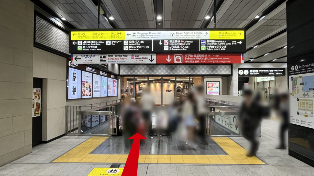 御堂筋線への連絡通路のエスカレーター付近　向かいには飲食店、頭上には大きな案内板がぶら下がっている