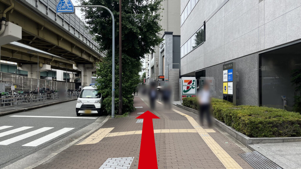 歩道橋を下に降りた時に見える風景　茶色のタイルが敷かれた歩道の左に横断歩道、右にセブンイレブンが写っている　左に高架下の道路がある