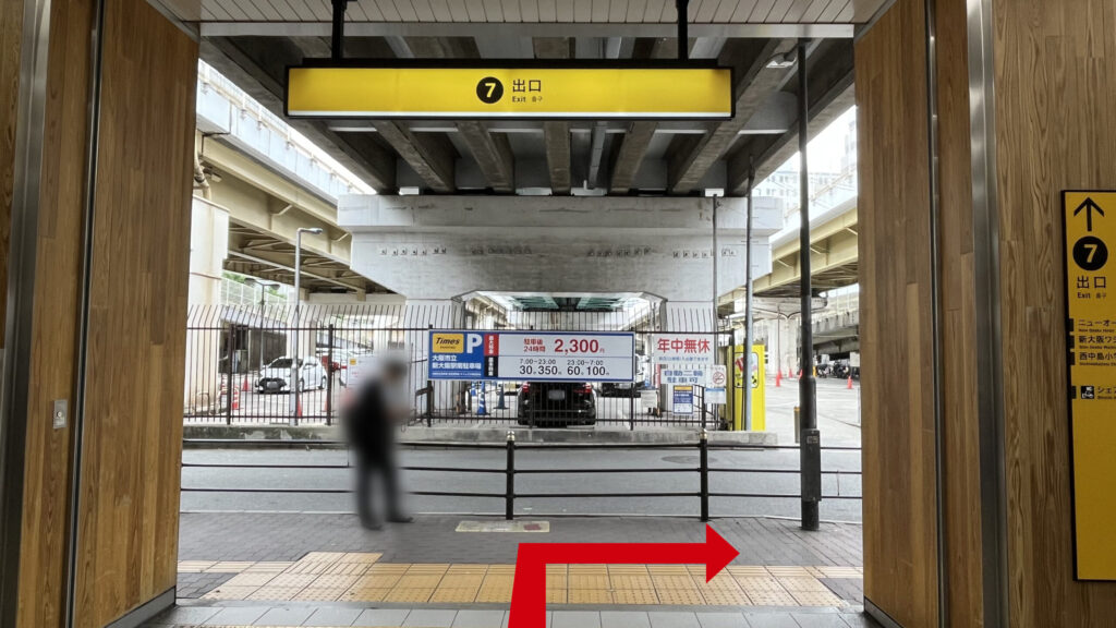７番出口付近の写真　頭上と右側に７番出口の黄色い看板、歩道の反対側に駐車場が広がっており、上には新御堂筋が走っている