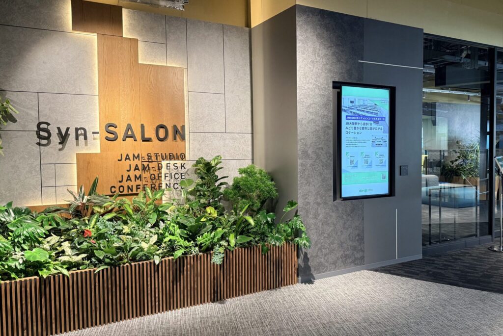 JAM BASEの入口付近　壁沿いに観葉植物が植えられており、その上に施設名が書かれている　右奥に入口がある
