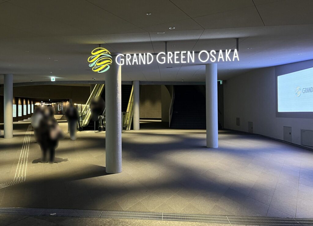 Signature グラングリーン ゲートタワーへ向かう、グラングリーンの連絡通路　頭上に「GRAND GREEN OSAKA」と書かれたネオンサインが掲示されており、円柱がいくつか建っている