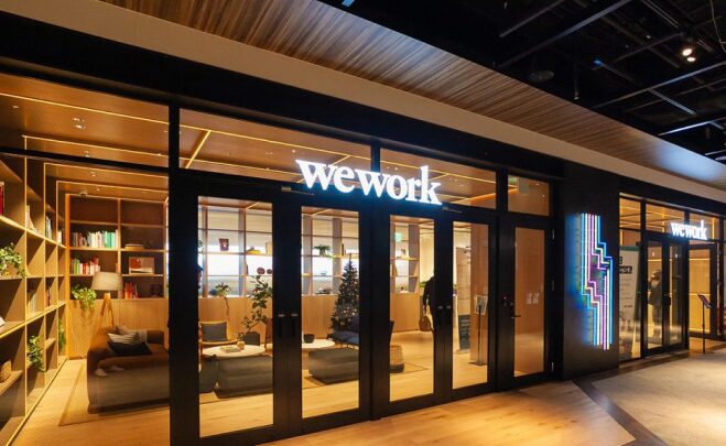 WeWork LINKS UMEDAの入口外観　暖かな照明とカフェのようにくつろげそうな受付周辺を外から見ている図　ガラス張りになっており、頭上に「wework」と書かれたネオンサインがある