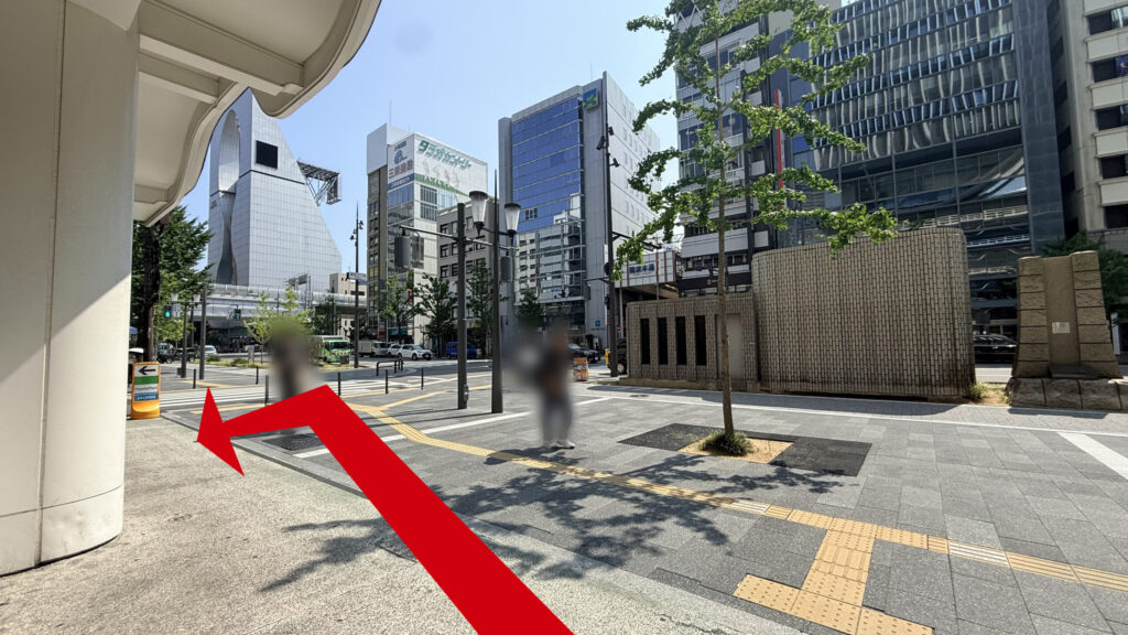 12番出口を出た地上付近の様子。左手に建物の柱があり、前方には大通りや周囲のビル群、木々が見える屋外スペースが広がっている。