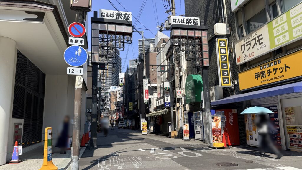 「なんば楽座」の路上。上部には大きな「なんば楽座」の看板や電線が交差し、街頭には「甲南チケット」などの店舗看板が並ぶ路地が伸びている。