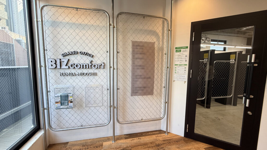 施設エントランス。フェンス状のパーティションに「SHARED OFFICE BIZcomfort NAMBA-MIDOSUJI」のロゴが掲げられ、右側にガラス扉の入口がある。