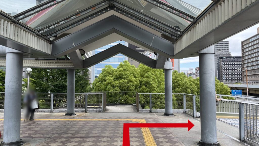 屋根とグレーの丸柱がある歩道橋の分岐地点。床は茶色のタイル模様で奥に青々とした街路樹が見える景色。