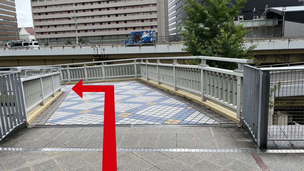 青と白の格子模様のタイル床が広がる歩道橋の曲がり角。周囲に白い金属製の手すりとフェンスが設置されている通路。