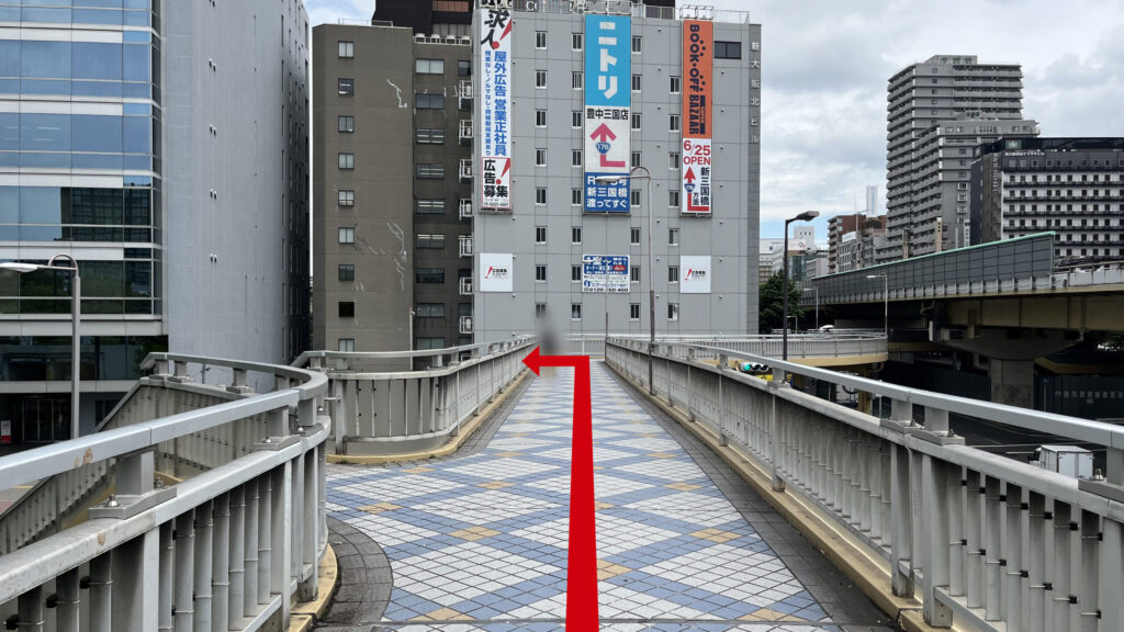 正面にニトリやBOOK-OFFの看板が掲げられたビルが見える直線の歩道橋。床面は青と白の格子模様になっている。