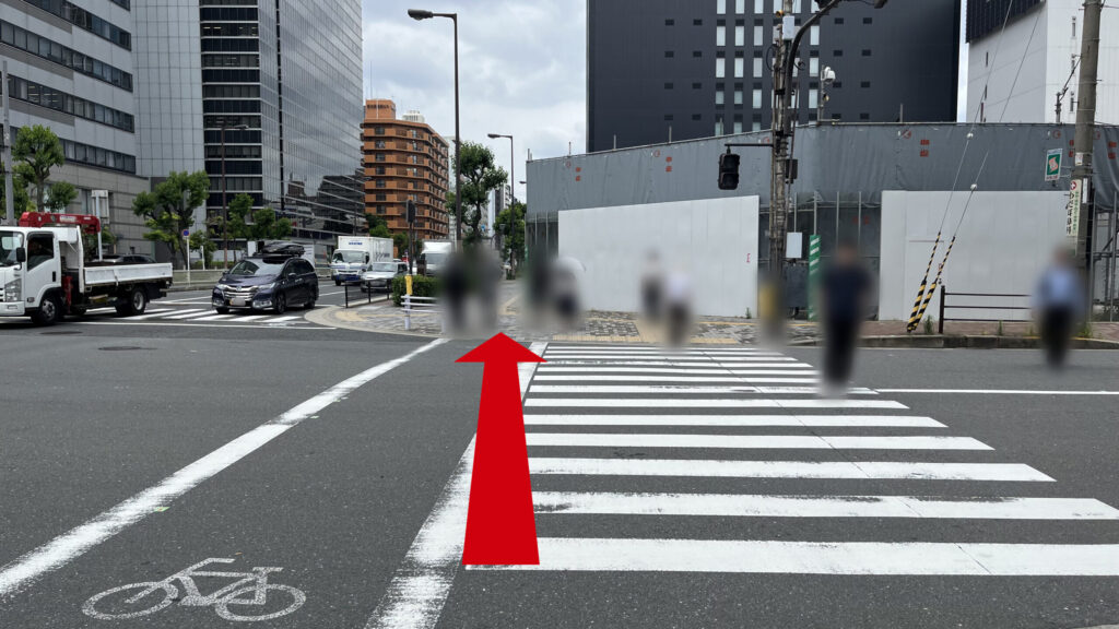 白線が描かれた横断歩道と対向の街並み。奥には黒いビルや工事用仮囲い壁が見える。