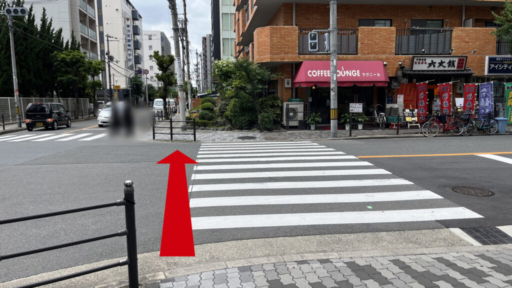 正面に横断歩道と1Fに喫茶店や飲食店のあるマンション、左側に道路が横断歩道と同方向に続いている様子 マンションやビルがたくさん建っている