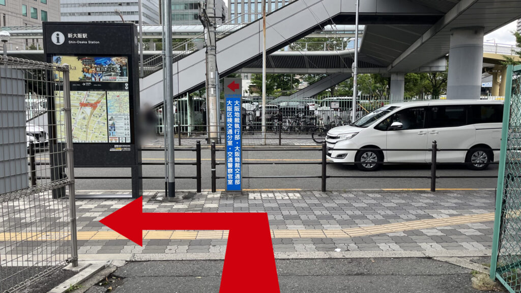 地上に下りた歩道の様子。左手には黒い「新大阪駅」の周辺案内図の看板があり、横に走る柵の向こうに車道があります。