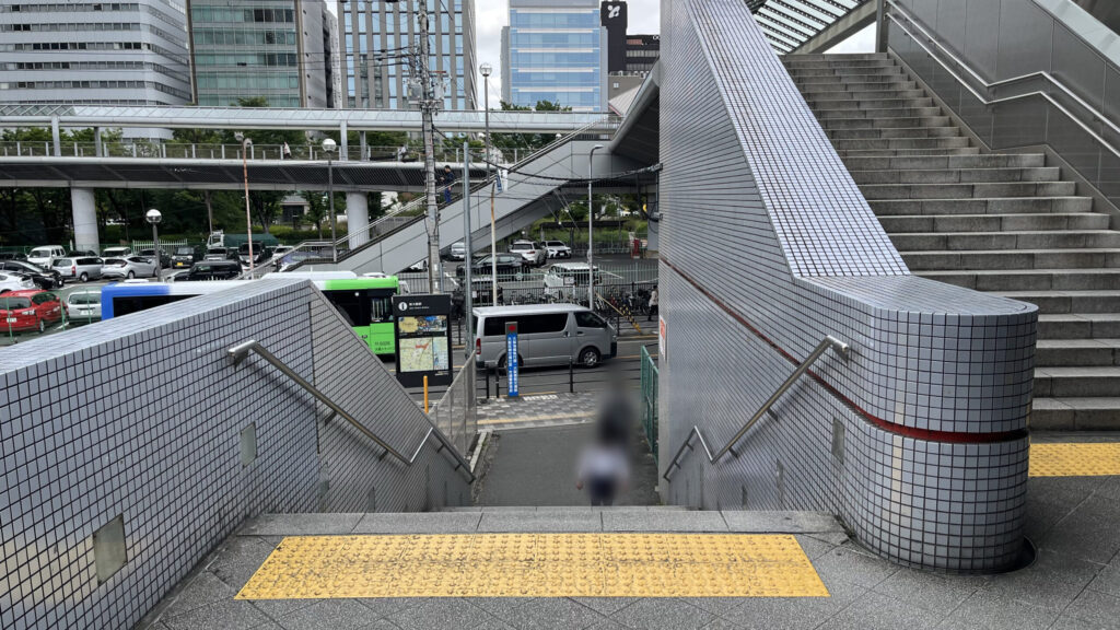 2階デッキから地上へと下りる、グレーのタイル張りの階段。階段の先には新大阪駅周辺の案内マップ看板や、道路を走る車が見えます。
