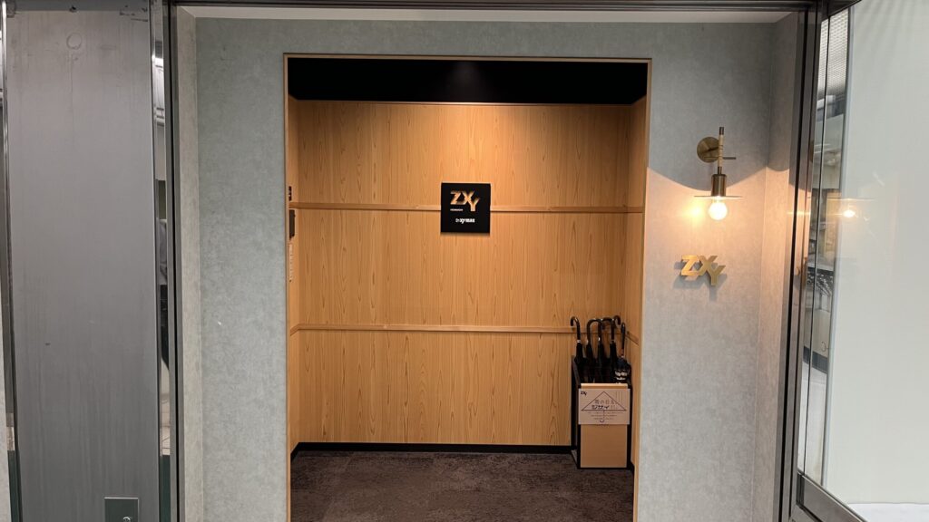 ZXY本町への行き方:ZXY本町エントランスを正面から撮った写真 奥まった木製の壁に「ZXY」と書かれた黒いパネルが貼られている